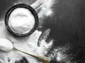 Biały proszek, prawdopodobnie soda na mrówki, rozsypany na czarnym tle. Obok miseczka i łyżka wypełnione proszkiem.