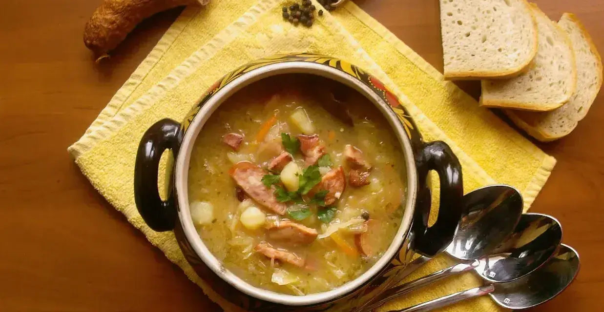 Aromaticzny kapuśniak z kiełbasą i ziemniakami, podany w ozdobnym naczyniu, obok kromek chleba i ziaren pieprzu.
