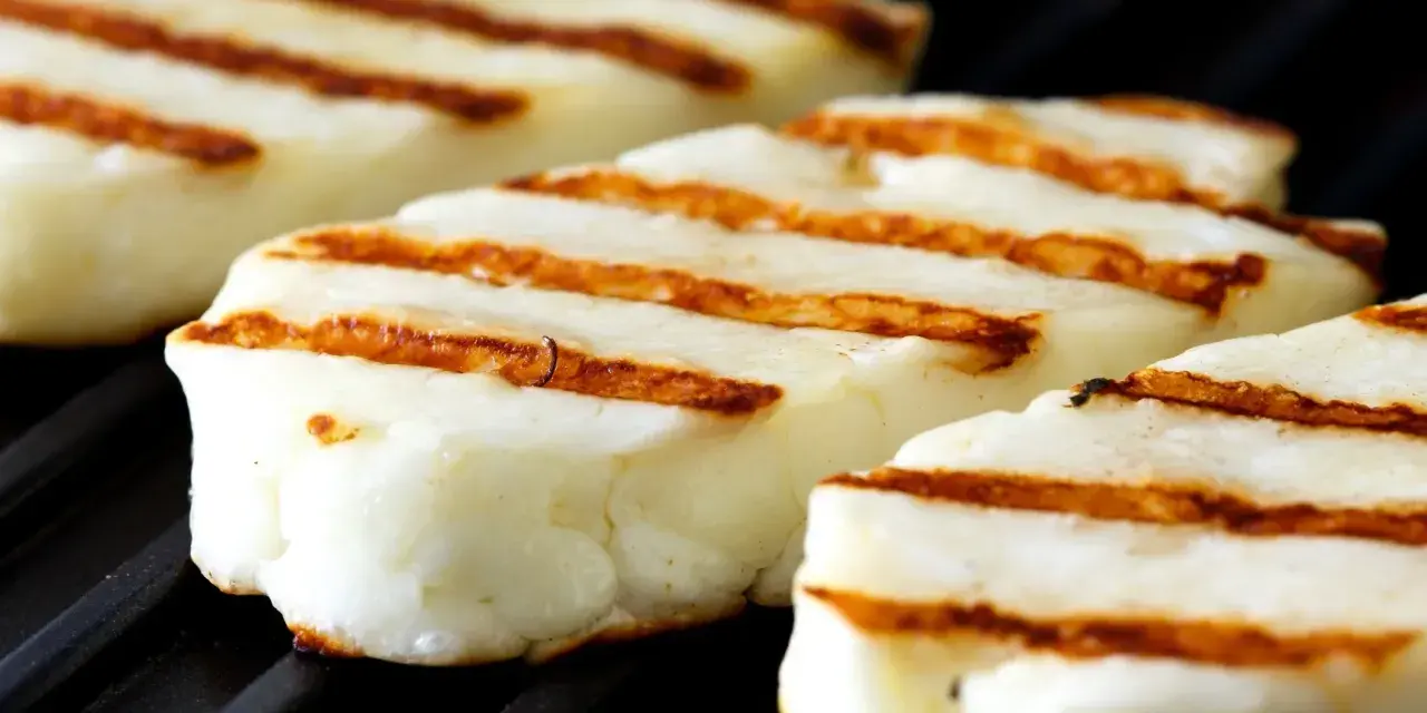 Trzy kawałki halloumi z grilla, z charakterystycznymi brązowymi paskami, leżą na czarnej powierzchni rusztu.