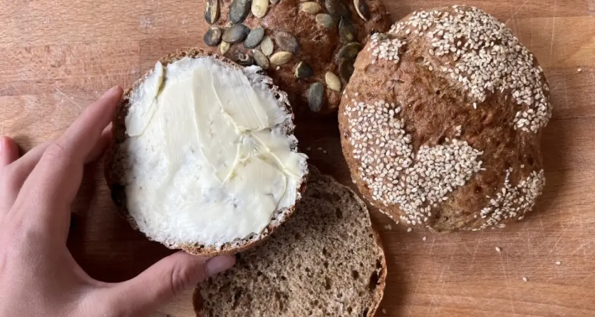 Ręka smaruje masłem bułkę z pestkami dyni, obok leżą inne bułki, w tym jedna z sezamem. Idealny chleb proteinowy na śniadanie.