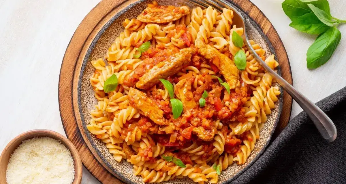 Pyszny makaron z kurczakiem w sosie pomidorowym, udekorowany bazylią. Obok miseczki z parmezanem i ziołami.