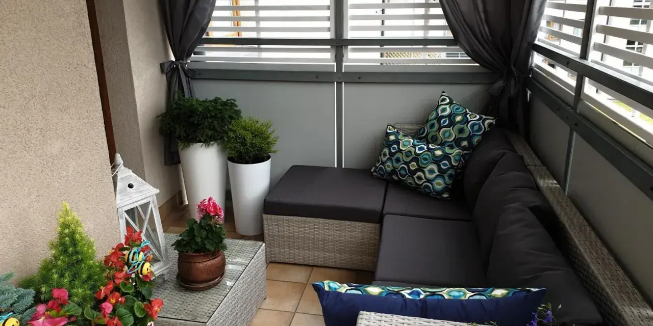 Przytulna zabudowa balkonu w bloku: rattanowy narożnik, poduszki w geometryczne wzory, rośliny i ozdobna latarenka tworzą relaksującą przestrzeń.