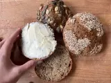 Ręka smaruje masłem bułkę z pestkami dyni, obok leżą inne bułki, w tym jedna z sezamem. Idealny chleb proteinowy na śniadanie.