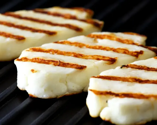 Trzy kawałki halloumi z grilla, z charakterystycznymi brązowymi paskami, leżą na czarnej powierzchni rusztu.