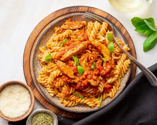 Pyszny makaron z kurczakiem w sosie pomidorowym, udekorowany bazylią. Obok miseczki z parmezanem i ziołami.