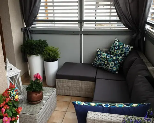 Przytulna zabudowa balkonu w bloku: rattanowy narożnik, poduszki w geometryczne wzory, rośliny i ozdobna latarenka tworzą relaksującą przestrzeń.