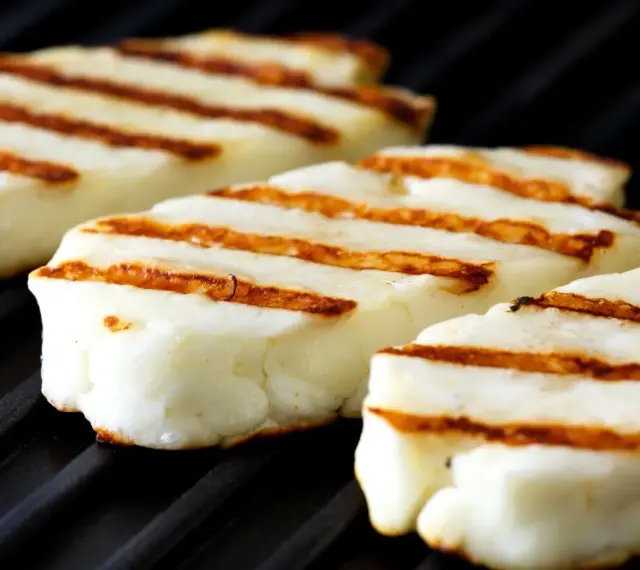 Trzy kawałki halloumi z grilla, z charakterystycznymi brązowymi paskami, leżą na czarnej powierzchni rusztu.