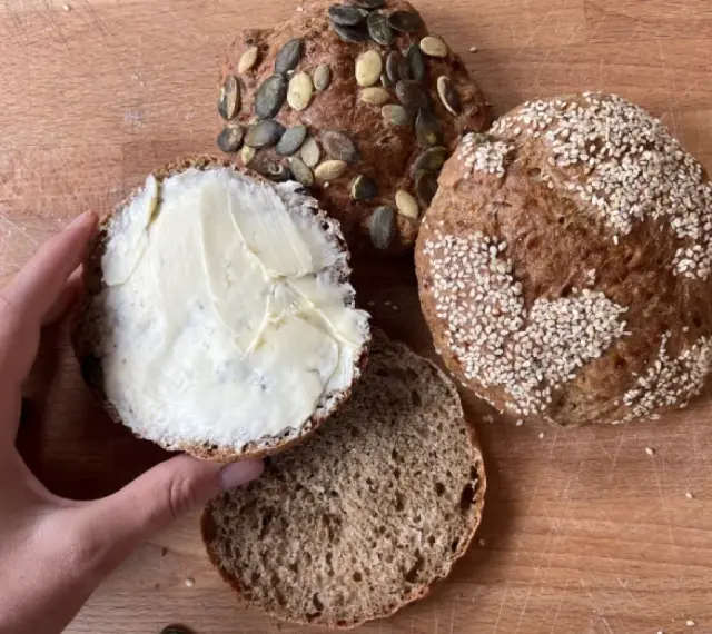 Ręka smaruje masłem bułkę z pestkami dyni, obok leżą inne bułki, w tym jedna z sezamem. Idealny chleb proteinowy na śniadanie.