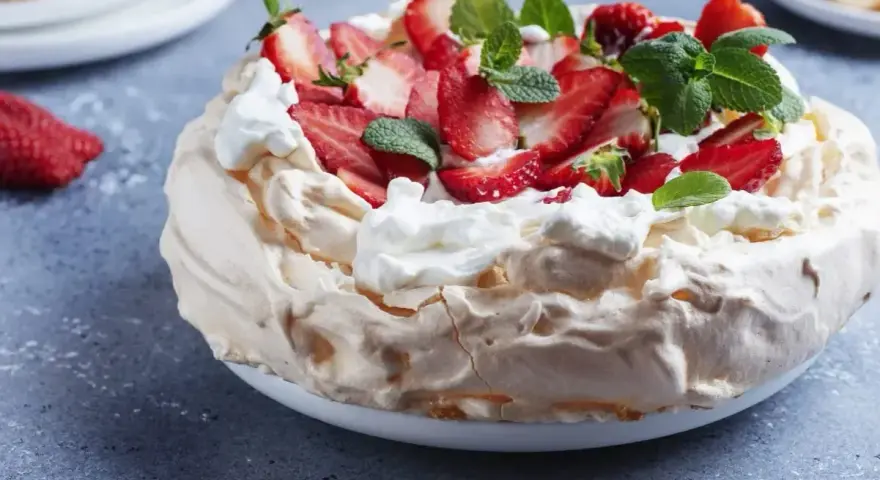Pyszny tort bezowy z mascarpone i bitą śmietaną, ozdobiony świeżymi truskawkami i miętą.