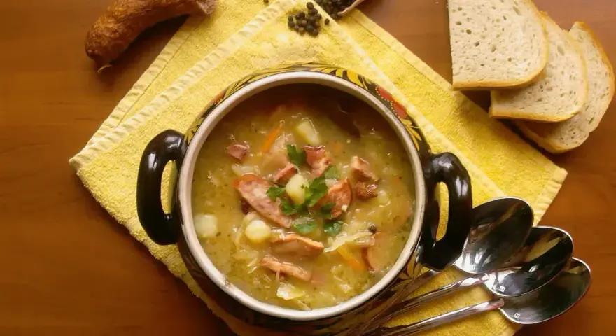 Aromaticzny kapuśniak z kiełbasą i ziemniakami, podany w ozdobnym naczyniu, obok kromek chleba i ziaren pieprzu.