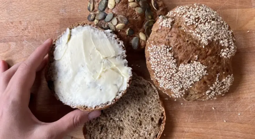 Ręka smaruje masłem bułkę z pestkami dyni, obok leżą inne bułki, w tym jedna z sezamem. Idealny chleb proteinowy na śniadanie.