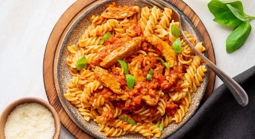 Pyszny makaron z kurczakiem w sosie pomidorowym, udekorowany bazylią. Obok miseczki z parmezanem i ziołami.