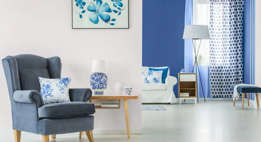Wygodny fotel w odcieniu **kolor chabrowy** i poduszka z motywem kwiatowym. Obok stolik z lampą i porcelaną.