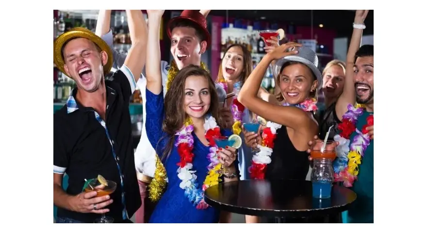 Impreza w stylu hawajskim: ludzie w kolorowych girlandach i kapeluszach tańczą i piją drinki.