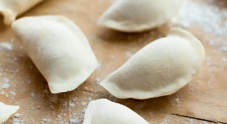 Surowe pierogi na drewnianej desce, przygotowane z ciasta na pierogi bez jajka.