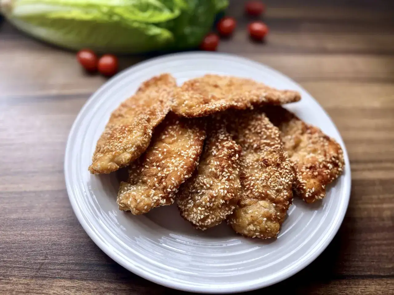 Chrupiący kotlet drobiowy w sezamie, ułożony na białym talerzu. W tle sałata i pomidorki.