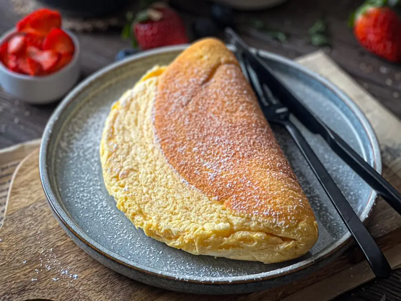 Puszysty omlet, lekko przyprószony cukrem pudrem, czeka na talerzu. Obok leżą czarne sztućce. W tle widać miseczkę z truskawkami.
