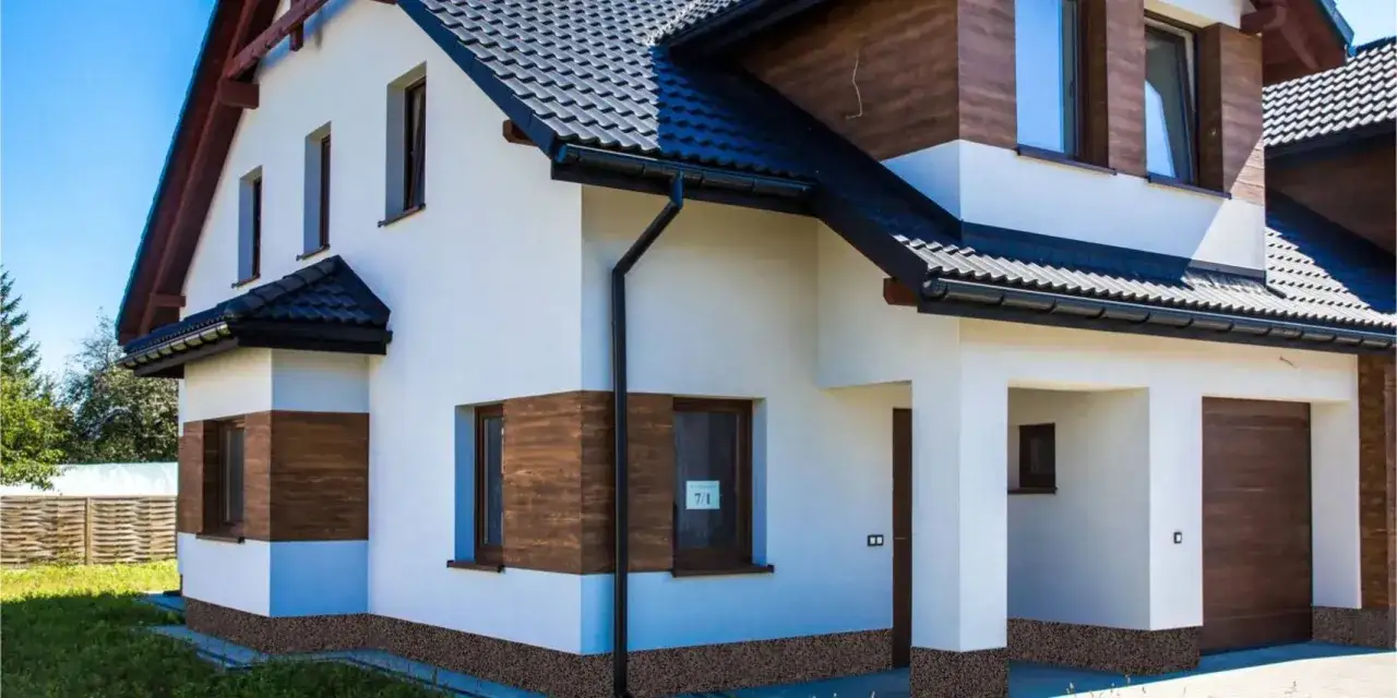 Nowoczesny dom z białymi ścianami i drewnianymi akcentami. Widoczny cokół na elewacji, ciemny dach i okna.