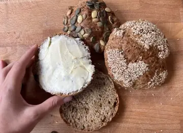 Ręka smaruje masłem bułkę z pestkami dyni, obok leżą inne bułki, w tym jedna z sezamem. Idealny chleb proteinowy na śniadanie.