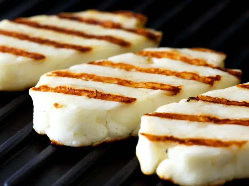 Trzy kawałki halloumi z grilla, z charakterystycznymi brązowymi paskami, leżą na czarnej powierzchni rusztu.