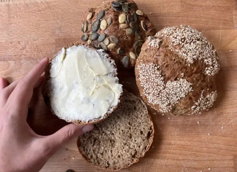 Ręka smaruje masłem bułkę z pestkami dyni, obok leżą inne bułki, w tym jedna z sezamem. Idealny chleb proteinowy na śniadanie.